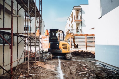 Mała koparka na gumowych gąsienicach pracująca na placu budowy pomiędzy budynkami mieszkalnymi.