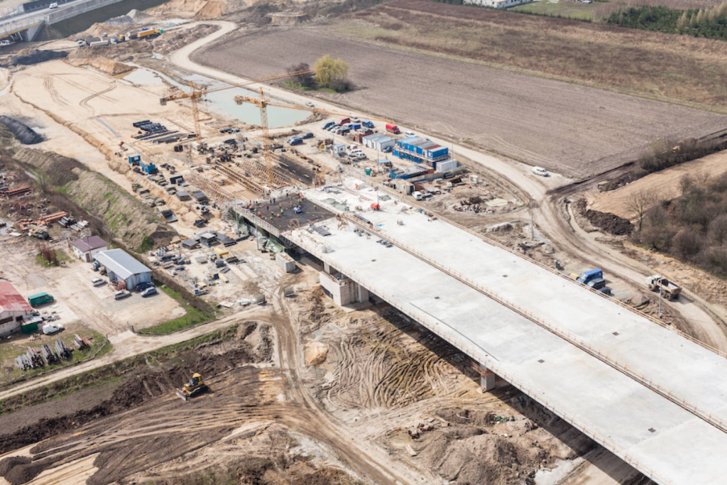 Widok z lotu ptaka na plac budowy mostu, z widocznymi dźwigami, maszynami budowlanymi i strukturami mostowymi.