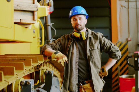 Pracownik budowlany w niebieskim kasku ochronnym i rękawicach roboczych, oparty o żółte podwozie maszyny budowlanej w warsztacie.