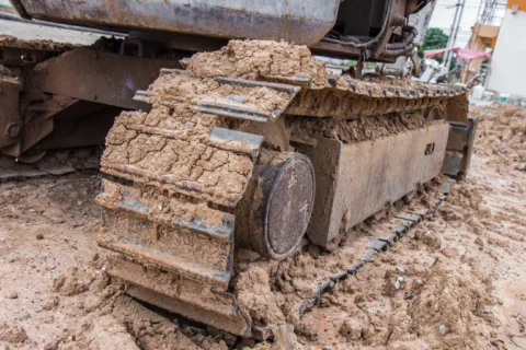 Podwozie koparki gąsienicowej z widocznymi metalowymi gąsienicami, rolką jezdną i kołem napinającym, pokrytymi zbitym błotem podczas pracy w grząskim terenie.